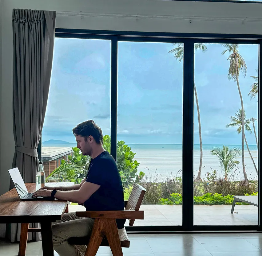 Ein Kollege arbeitet an einem Laptop an einem Holztisch in einem Raum mit großer Fensterfront. Draußen sind Strand, Meer und Palmen zu sehen. Die Szene zeigt Remote-Arbeit in einer ruhigen Umgebung mit Blick auf die Natur.